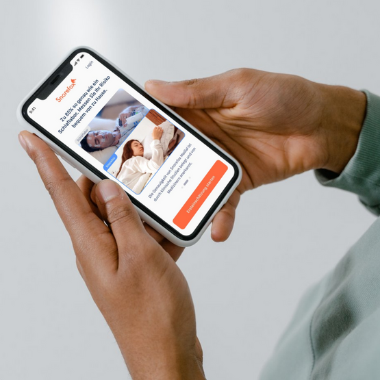 Zwei Hände halten ein Smartphone: man sieht auf dem Smartphone ein Schlaflabor und wie man mit der Snorefox M App neben sich schläft 